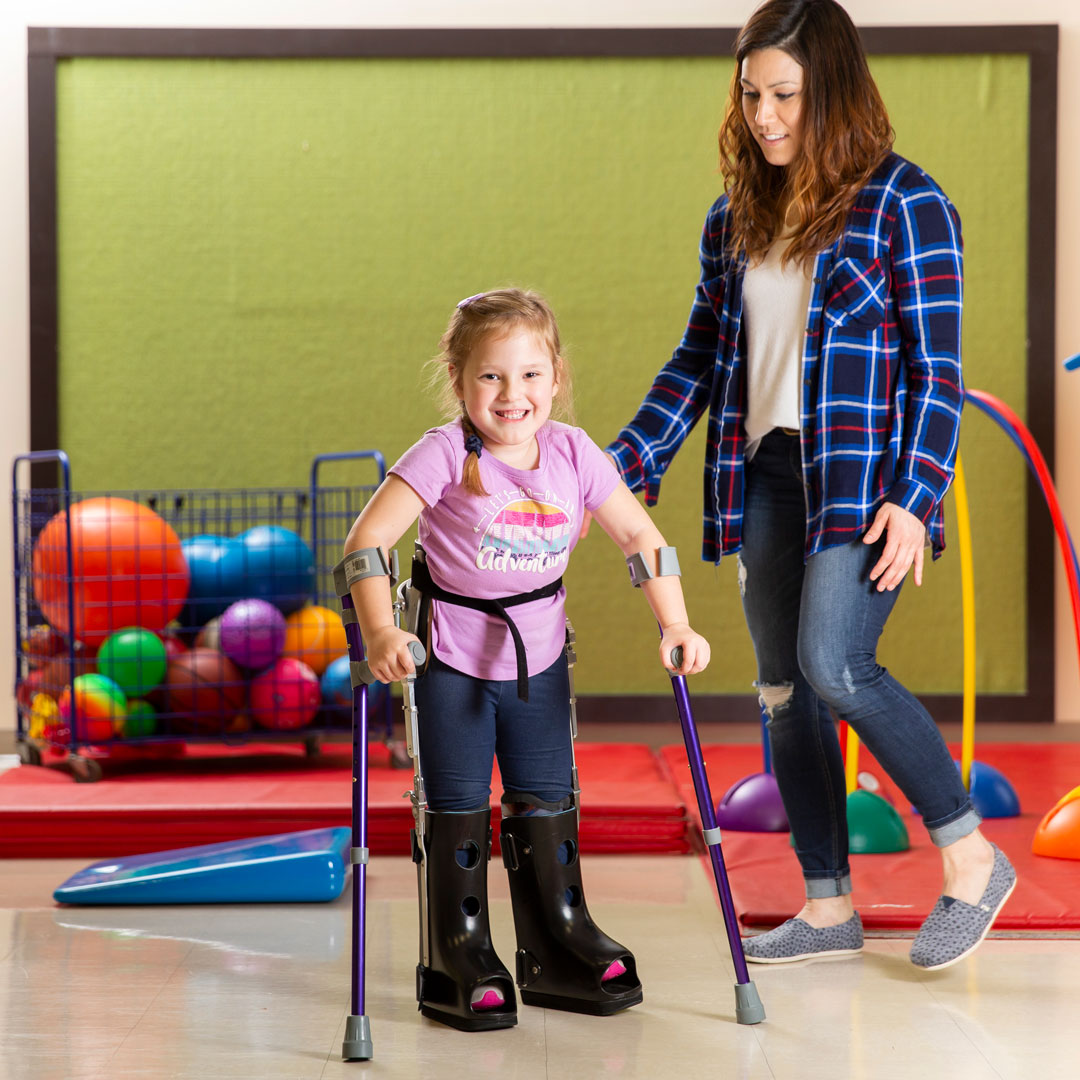 Image of little girl, Willow, standing on a matt in a therapeutic room with leg braces. Mom is standing next to her smiling.